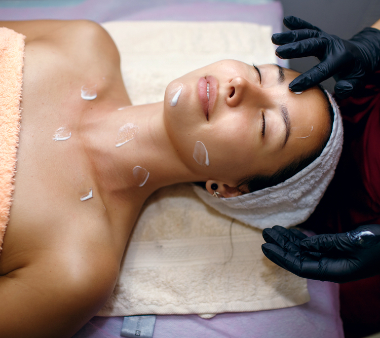 A beautiful brunette is undergoing procedures at the spa, a master beautician applies a moisturizer to her face. Facial skin care concept, professional lifting, free space for text